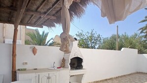 En udendørs patio med stenbelægning, grill i hvid farve og et overdækket siddeområde.