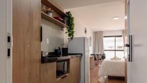 A compact kitchen with wooden cabinets, a built-in oven, and a refrigerator. There is a dining area with a table and chairs, and a bed with a white bedspread in the background. - Cozy Apartment Close to the Center Pgo309 (Poços de Caldas)