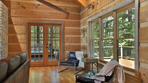 A log cabin interior with a wooden floor, a leather couch, a dining table with chairs, and large windows offering a view of the outdoors.