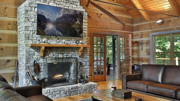 A cozy cabin interior with a stone fireplace, a leather sofa, and a large window offering a view of the outdoors.