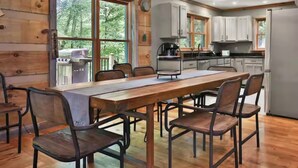 A wooden interior with a dining table and chairs, a kitchen area with cabinets and appliances, and large windows offering a view of trees.