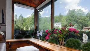 Interior de madera con un banco, otomanes y vistas a zonas verdes y una casa a través de amplios ventanales.