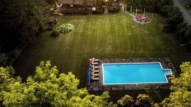 Una casa grande rodeada de un bosque frondoso, con piscina y zona de estar al aire libre.