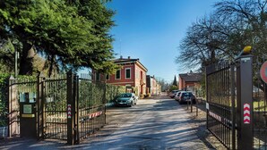 A gated entrance with a red building in the background, a paved driveway, and parked cars. - Italian flat - Verona's River (Verona)