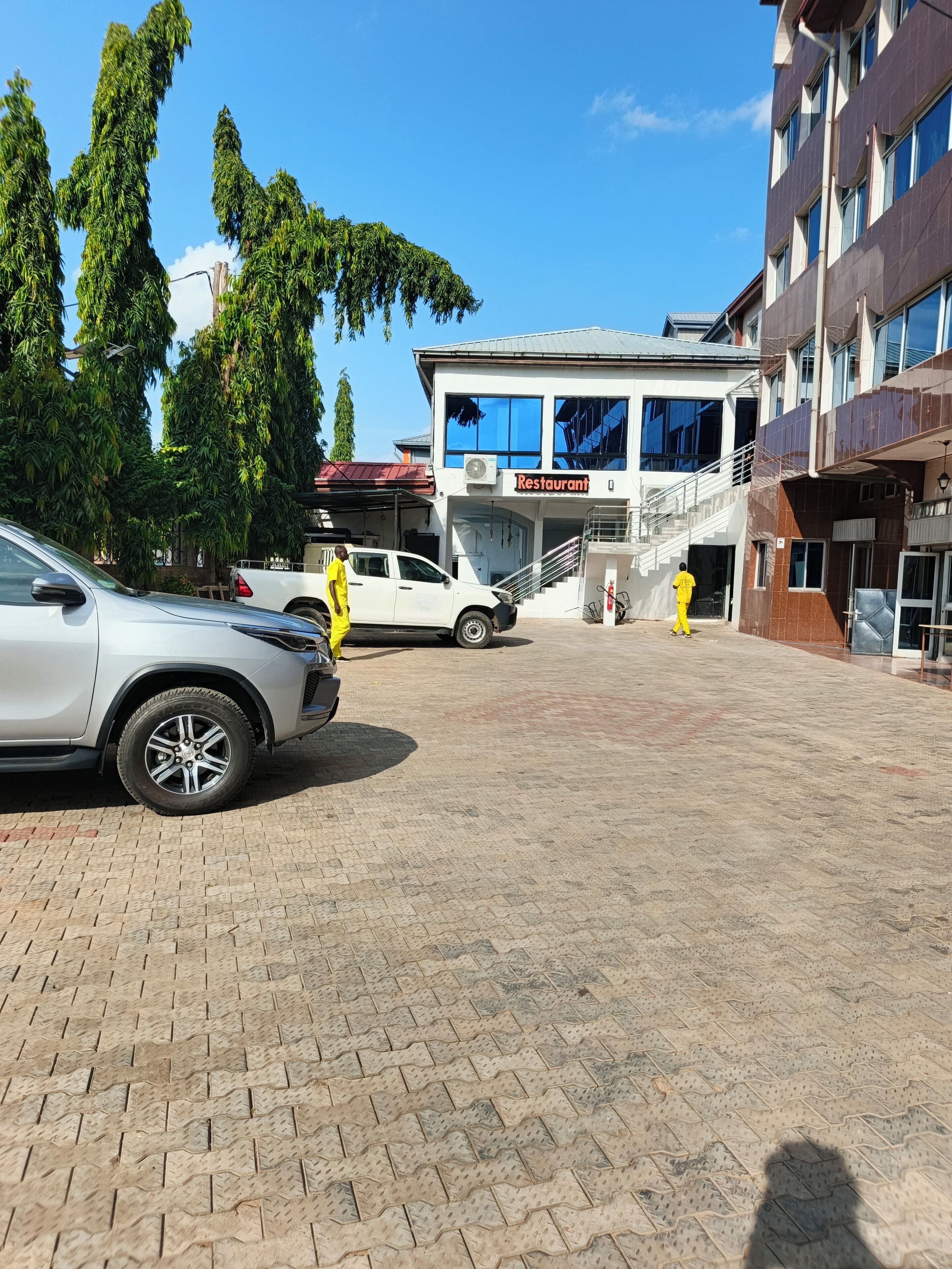 Un área pavimentada con un edificio que indica 'Restaurant' al fondo, un coche blanco estacionado a la izquierda y una persona vestida de amarillo caminando hacia el edificio.