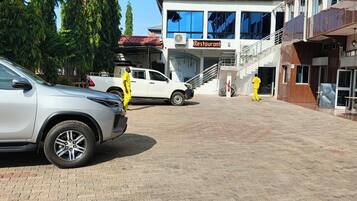 Un área pavimentada con un edificio que indica 'Restaurant' al fondo, un coche blanco estacionado a la izquierda y una persona vestida de amarillo caminando hacia el edificio.