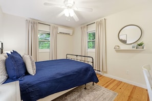 A bedroom with a bed, a ceiling fan, an air conditioning unit, a window with curtains, a mirror, and a small shelf with a plant.