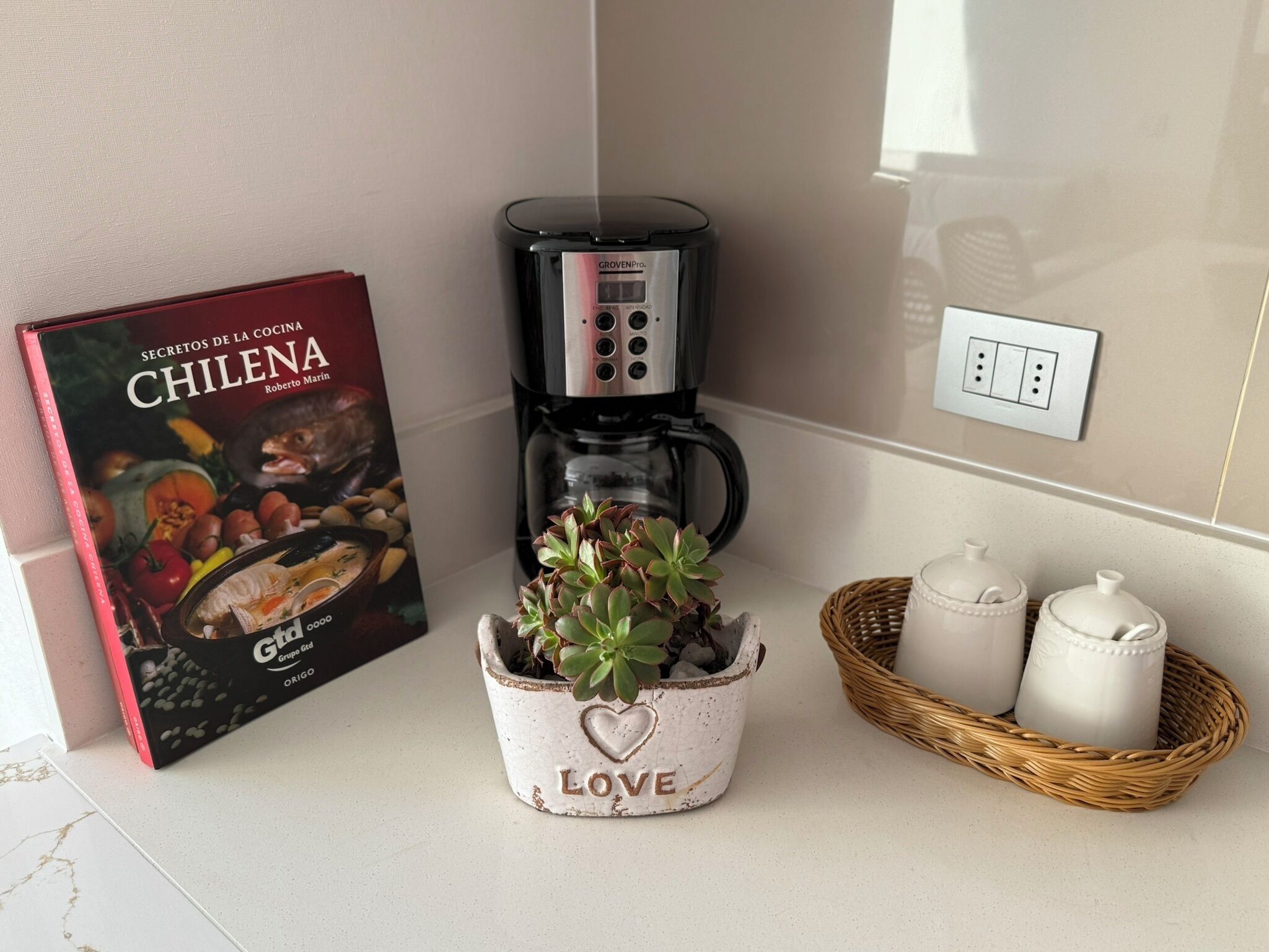 En køkkenbordplade med en kaffemaskine, en bog med titlen 'CHILENA', en dekorativ plantekasse med et hjerte og to keramiske beholdere.