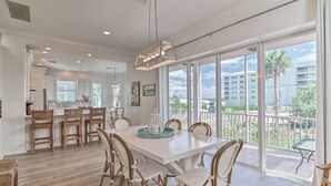 A spacious dining area with a white table and wicker chairs, large windows with a view of buildings and palm trees, and a modern kitchen in the background.