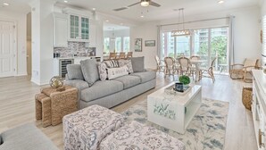 A spacious living room with a grey sofa, a coffee table, and wicker side tables. There is a dining area with a white table and wicker chairs. The room has a ceiling fan, large windows, and a view of greenery outside.