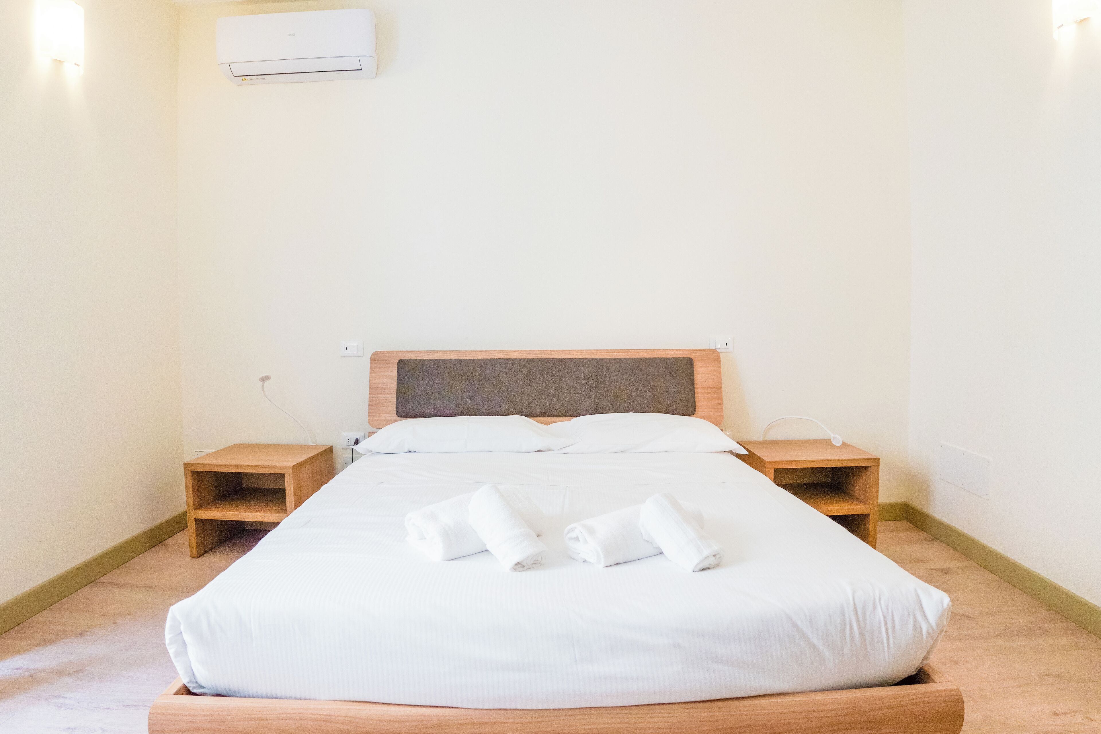 A neatly made bed with white linens and rolled towels, wooden headboard, bedside tables, and a wall-mounted air conditioner.