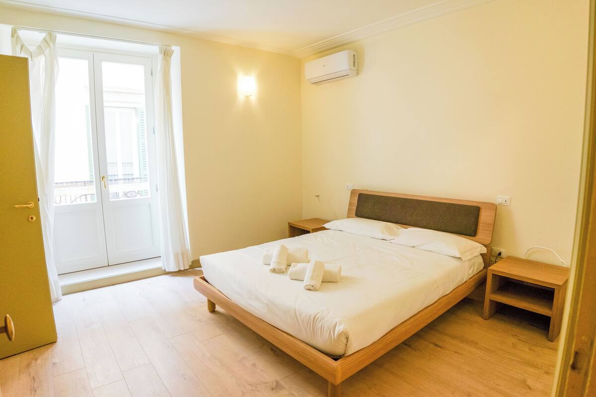 A bedroom with a wooden bed, a nightstand, a small wooden table, a wardrobe, and a sliding glass door to a balcony.