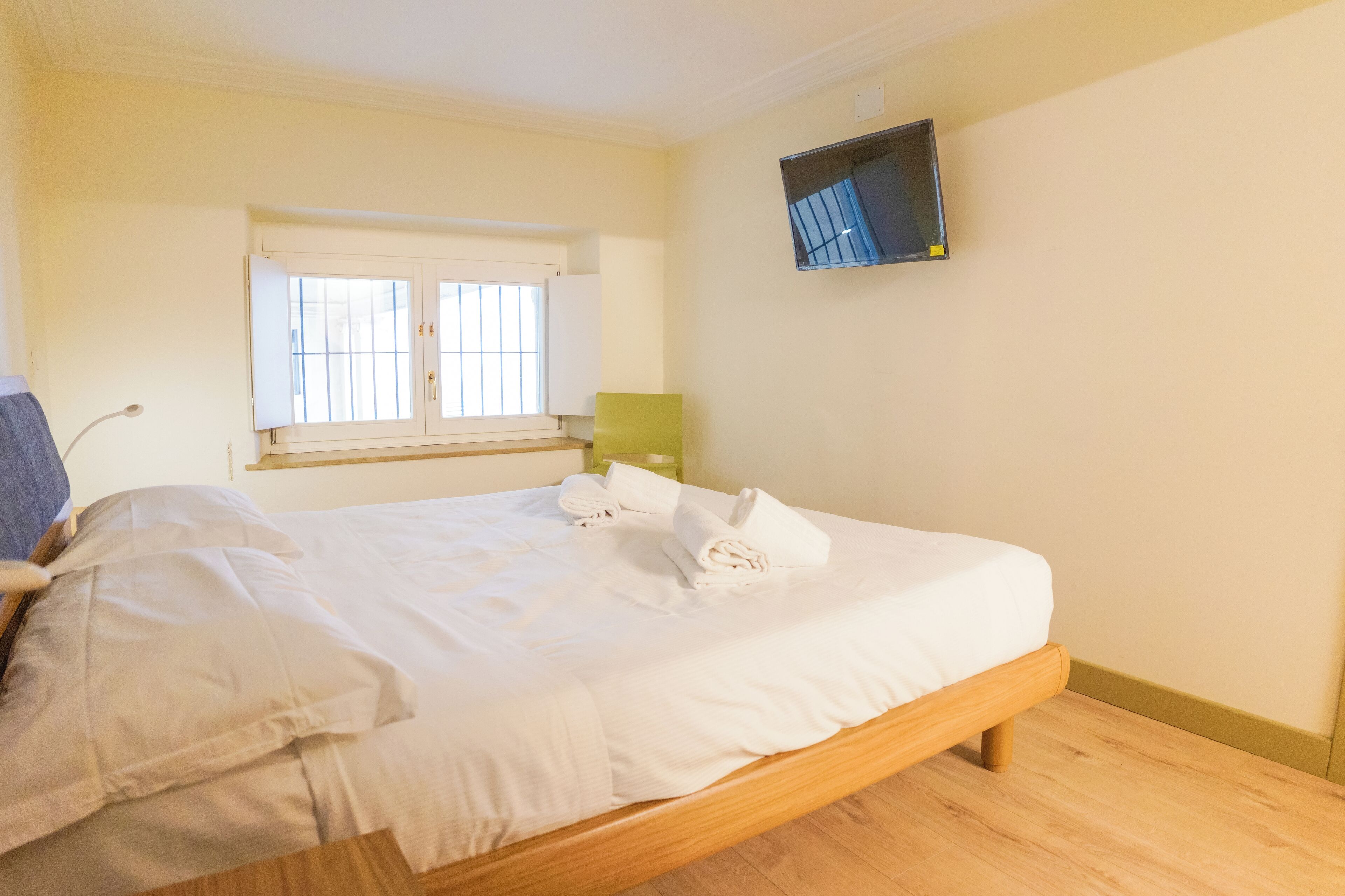 Un dormitorio con cama de madera, una ventana con persianas, un televisor de pantalla plana en la pared y una mesita de noche con lámpara.