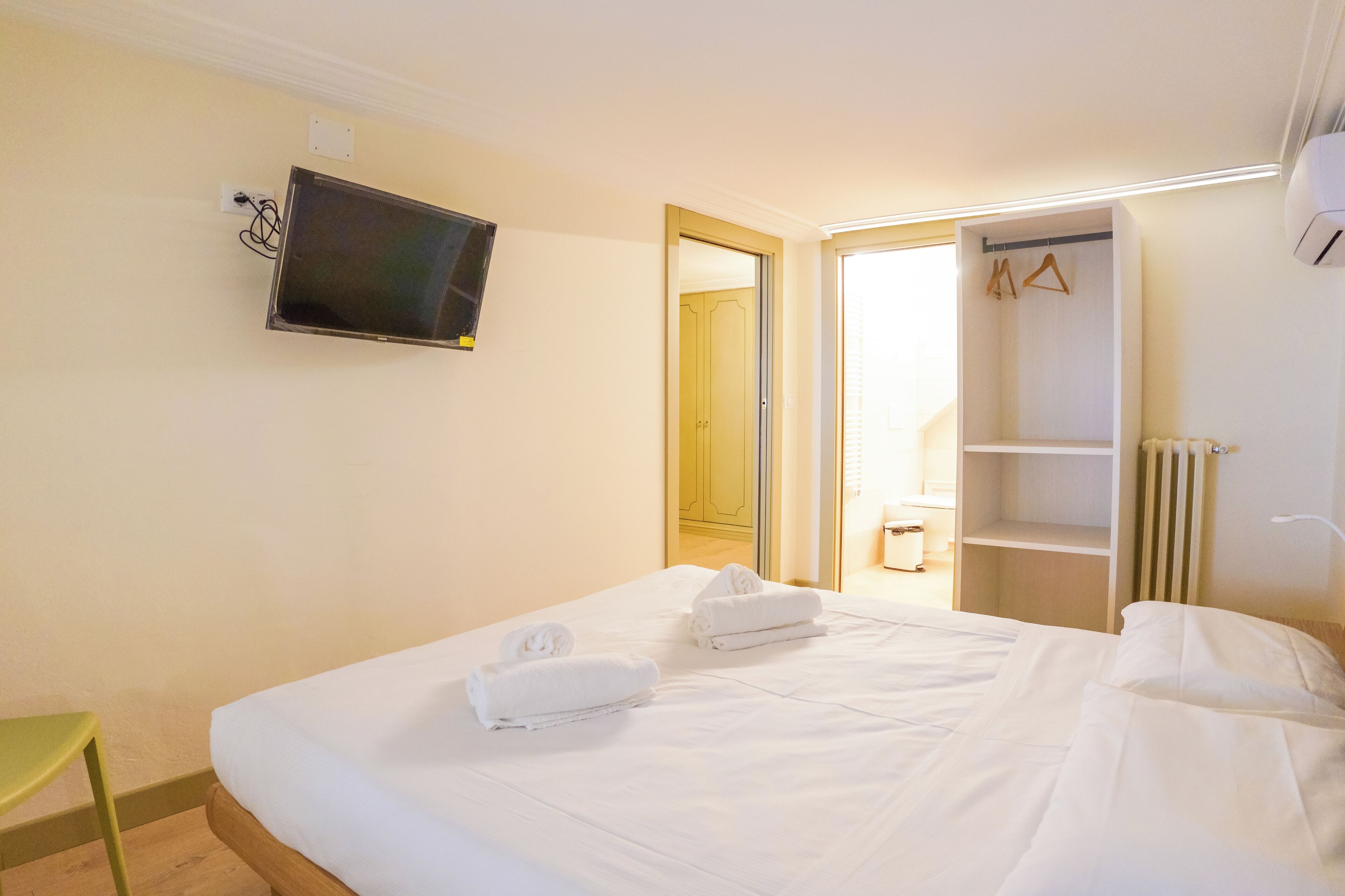 A hotel room with a bed, two towels, a television mounted on the wall, and a walk-in closet with a radiator.