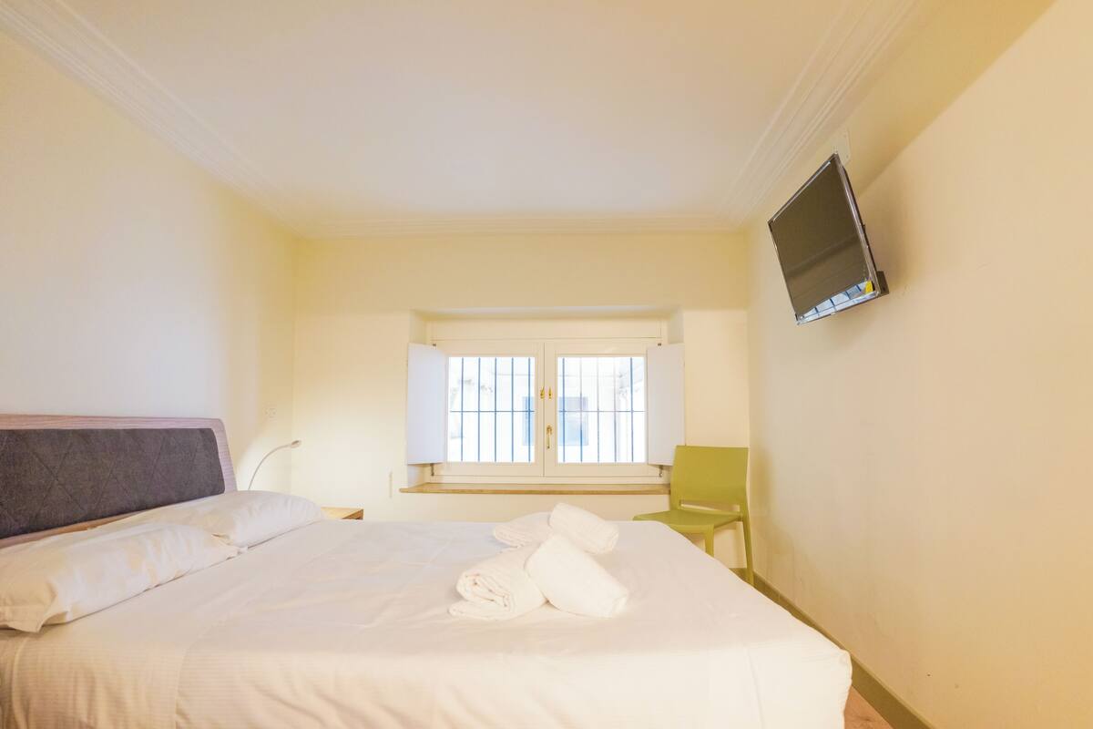A hotel room with a bed, a window with blinds, a chair, and a flat-screen TV mounted on the wall.