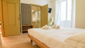 Ein Hotelzimmer mit Holzboden, einem Bett mit weißen Leinentüchern, einem Badezimmer mit Glasdusche und einem an der Wand befestigten Fernseher.