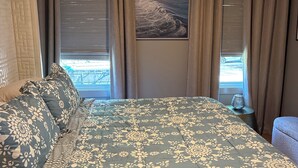 A bedroom with a large bed, a headboard, a window with curtains, a wall-mounted artwork, and a bedside table with a lamp.
