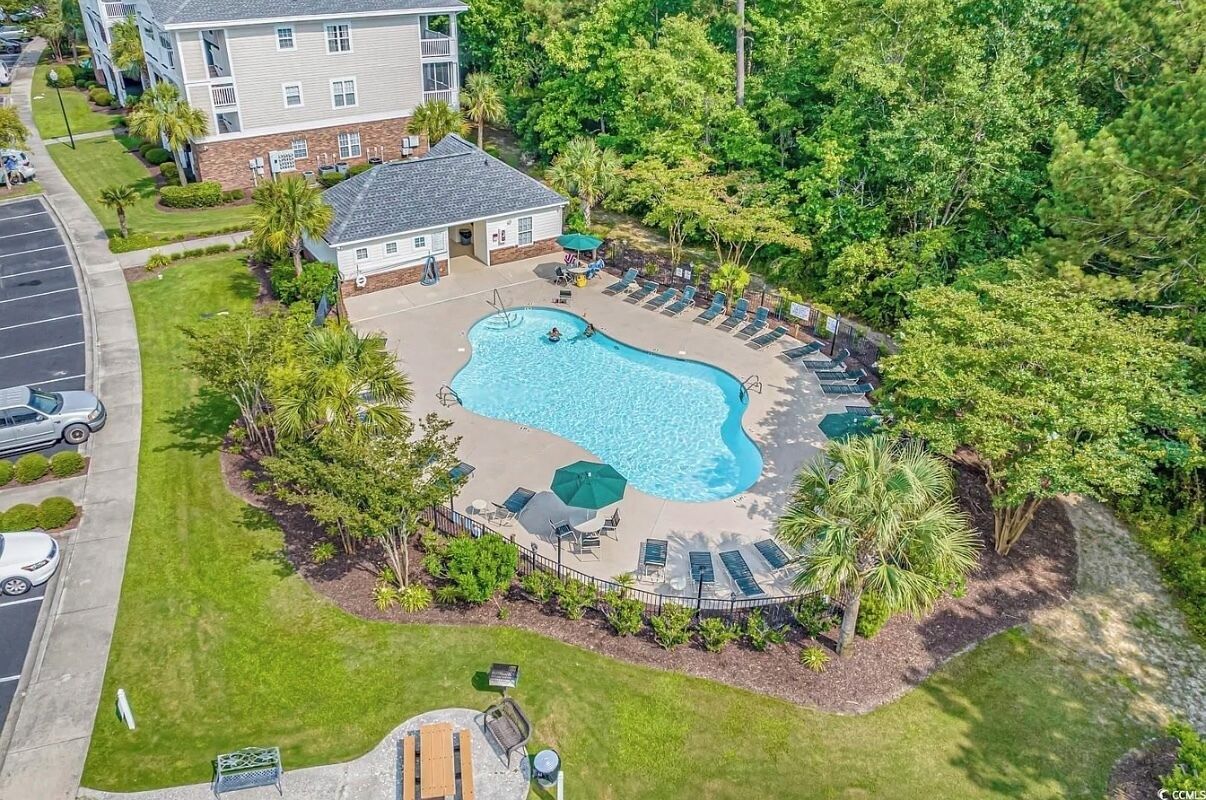 An aerial view of a hotel with a swimming pool surrounded by lounge chairs and umbrellas, set in a lush green environment.