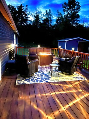 A wooden deck with outdoor furniture, a fire pit, and a view of the surrounding trees and sky. - Cozy 2BR cottage near the Harbor, beach, hunting and fishing! (Henderson)
