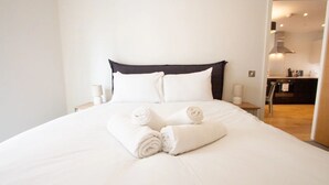 A neatly made bed with white linens and rolled towels, a dark headboard, and bedside lamps. - The Slate Yard (Salford)