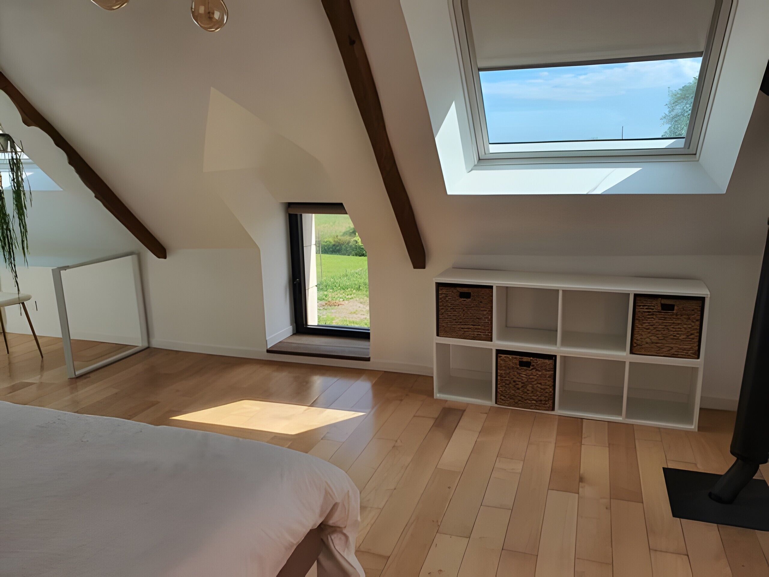 A bedroom with a sloped ceiling, a skylight, a bed, a white console table, and a view of the outdoors.