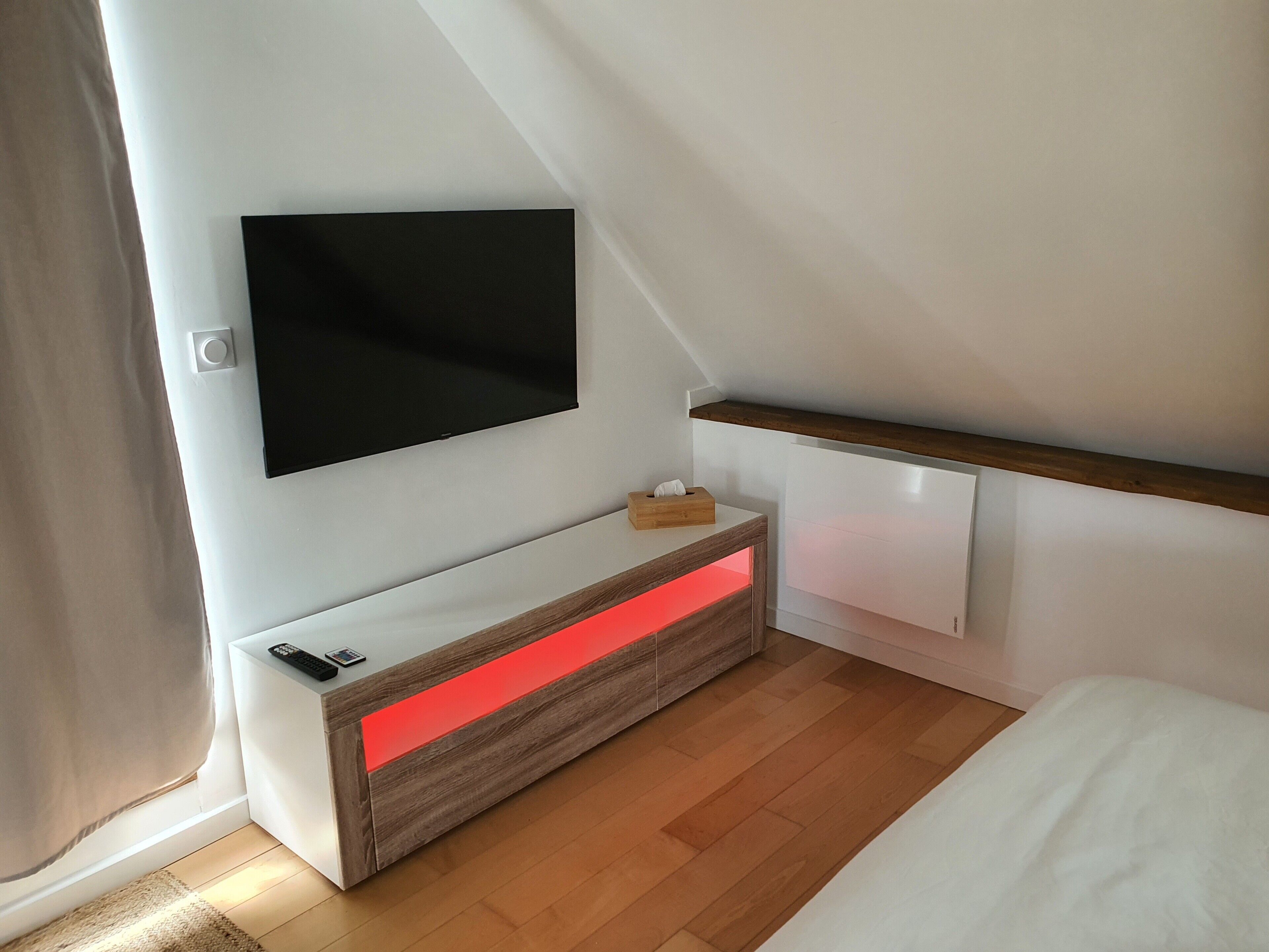 Een moderne slaapkamer met een aan de muur bevestigde televisie, een houten nachtkastje met een rode lamp, en een bed met wit beddengoed.