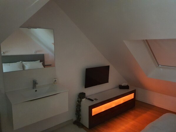 A bedroom with a sloped ceiling, a bed, a bedside table, a mirror, a TV, and a wall-mounted shelf.