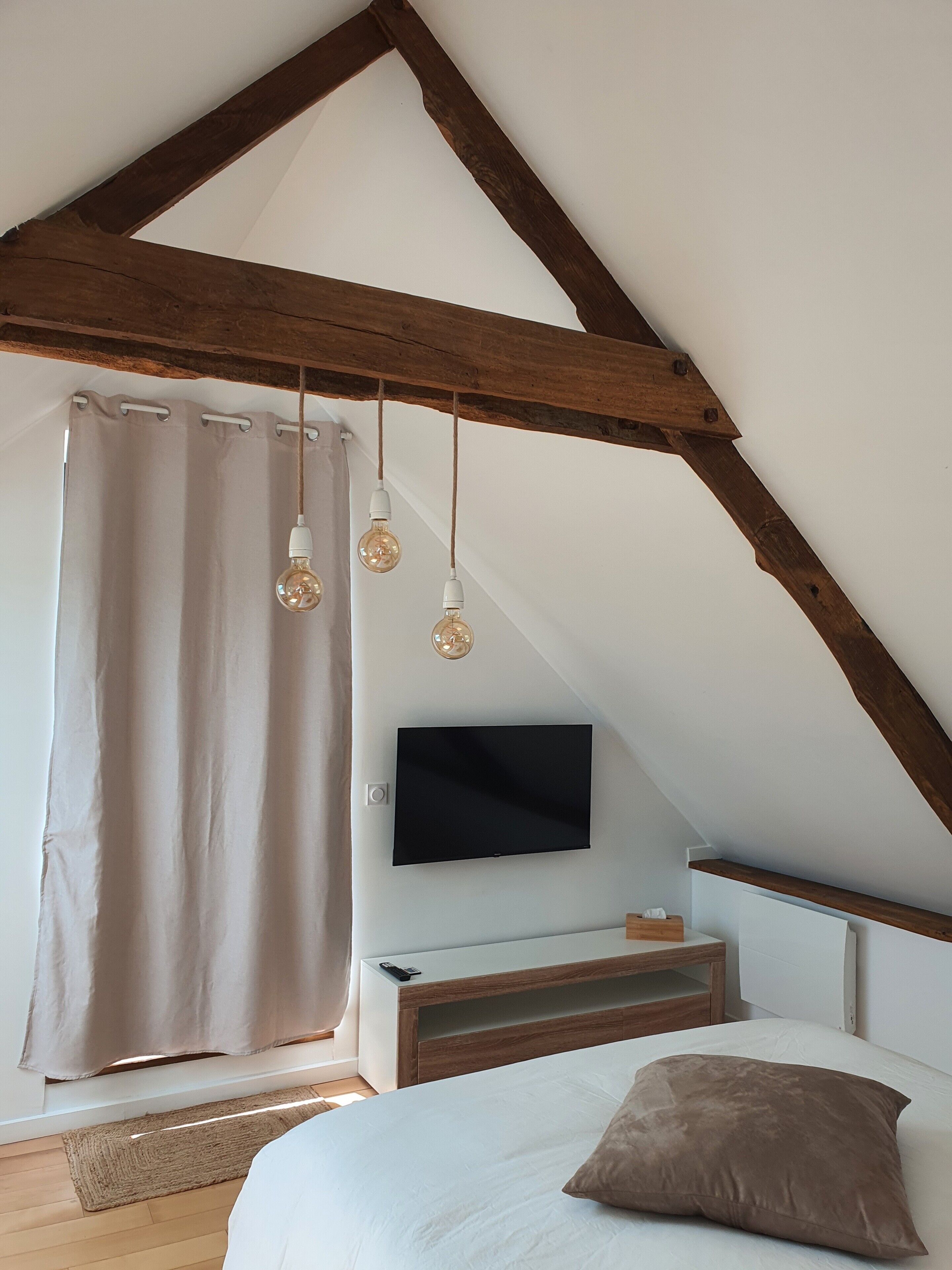 A bedroom with a sloped ceiling, a bed, a TV, and hanging light fixtures.