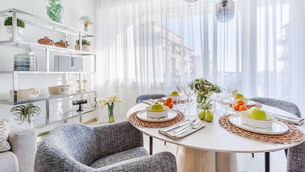 A dining area with a round table set for four, surrounded by grey upholstered chairs. A white dining table with a woven placemat is set with plates, glasses, and a centerpiece of green apples and pears. The room features a white wall with a glass display shelf holding decorative items and plants. - Versilian Tuscan coast escape (Viareggio)