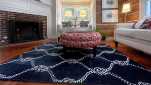 A living room with a fireplace, a patterned ottoman, a sofa, and a rug. - The Snuggery Inn & Suites Captain`s Quarters (Saint Michaels)