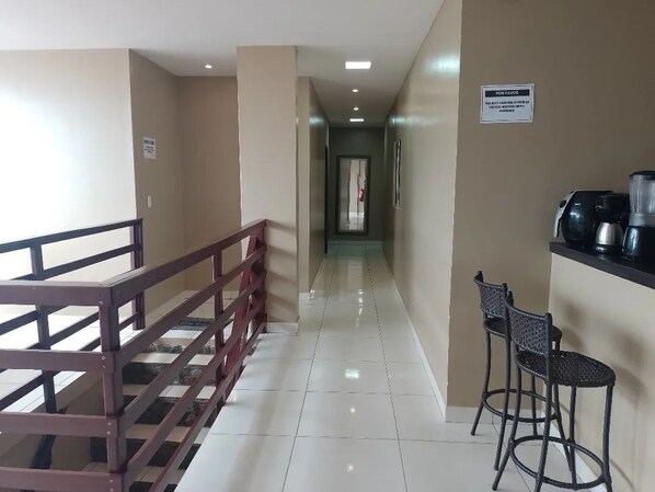 A corridor with a metal railing on the left, a coffee machine and bar stools on the right, and a sign on the wall.