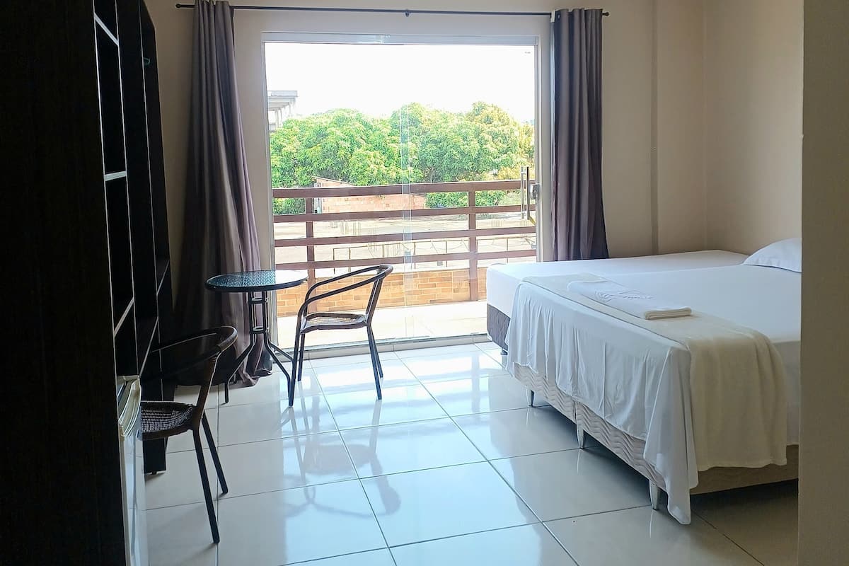 A hotel room with a bed, a small table with chairs, a view of greenery outside, and a balcony.