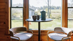 Ein Holzblockhaus-Interieur mit zwei stahlummantelten Stühlen, einem kleinen runden Tisch und Blick auf eine ländliche Landschaft durch große Fenster.