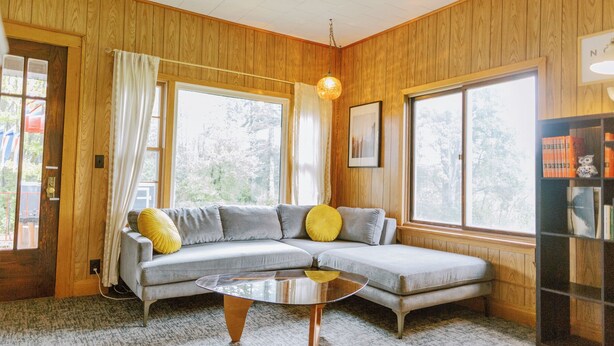 Ein gemütliches Zimmer mit einem grauen Ecksofa, einem Glastisch und einem Holzregal.