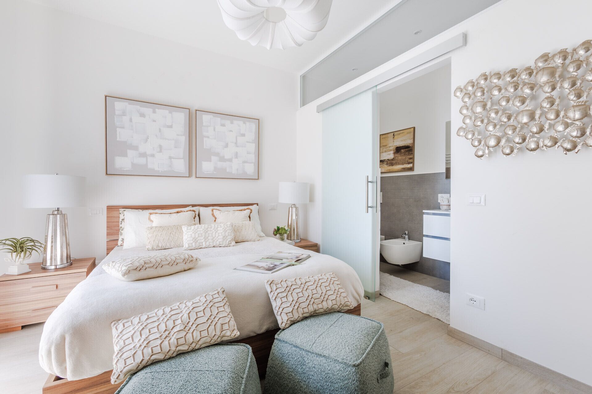 Une chambre moderne avec un grand lit, deux tables de chevet, une petite table d’appoint, une lampe fixée au mur et un élément décoratif mural.