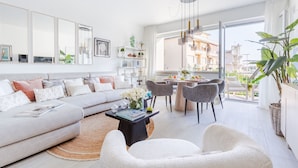 Un salon moderne avec un canapé, une table basse et des fauteuils. On y trouve également un coin repas avec une table et des chaises. La pièce est dotée de grandes fenêtres et offre une vue sur les bâtiments environnants.