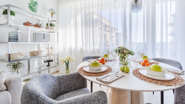 Un coin repas avec une table ronde dressée pour quatre personnes, entourée de chaises à revêtement gris. Une table à manger blanche, surmontée d’un set de table en fibres tressées, est mise en place avec des assiettes, des verres et un centre de table composé de pommes et de poires vertes. La pièce est agrémentée d’un mur blanc sur lequel est fixée une étagère en verre où sont exposés des objets décoratifs et des plantes.