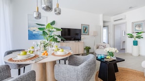 Un salon moderne avec un coin repas, comprenant une table ronde, des chaises recouvertes d’un tissu gris et un téléviseur à écran plat.