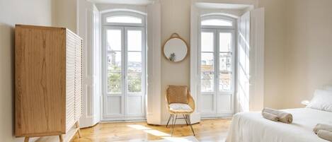 Ein Schlafzimmer mit Holzschrank, Korbsessel und Bett mit weißer Bettwäsche, sowie ein Blick durch große Fenster.