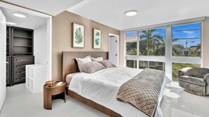 Ein modernes Schlafzimmer mit einem großen Bett, einem Nachttisch, einem Sessel, einem Kleiderschrank und einem Fenster mit Blick auf Palmen.
