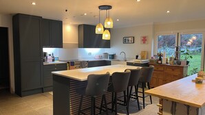 Une cuisine moderne avec des meubles de couleur sombre, un îlot central doté de tabourets de bar, un éclairage suspendu et une vue sur l’extérieur grâce à de grandes fenêtres.