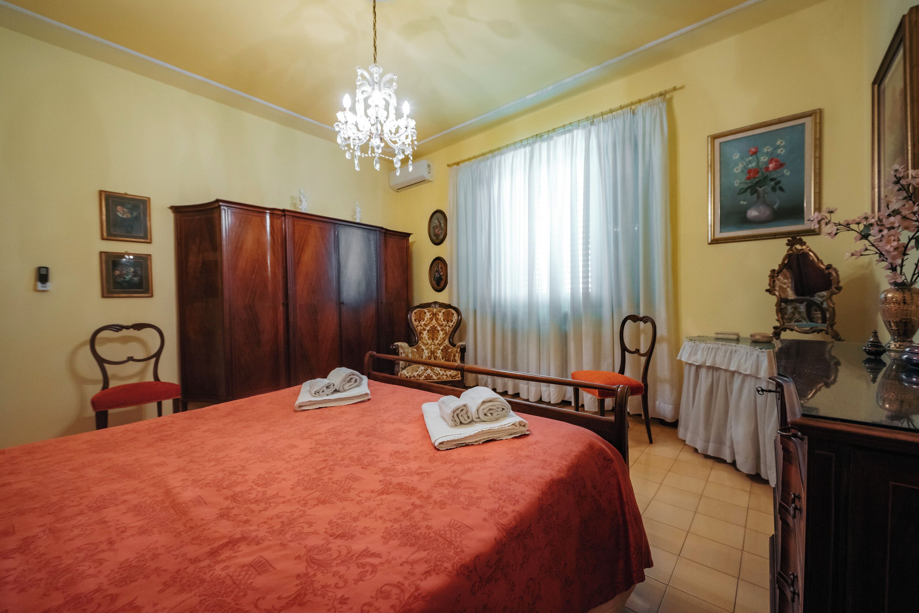 A room with a bed, a wooden wardrobe, a chandelier, a table with a vase, and a painting on the wall.