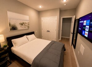 Un dormitorio con una cama, un televisor montado en la pared y una puerta que da a otra habitación.