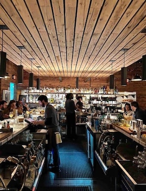 Un bar con vigas de madera en el techo, taburetes de bar y un bartender preparando bebidas.