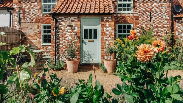 A two-story house with a red-tiled roof and brick walls. There are two windows on the front, a door, and a small garden with potted plants and flowers in front of the house.