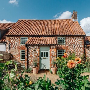 A two-story house with a red-tiled roof and brick walls. There are two windows on the front, a door, and a small garden with potted plants and flowers in front of the house. - Characterful period cottage close to beaches and local eateries. Dog-friendly! (Stanhoe)