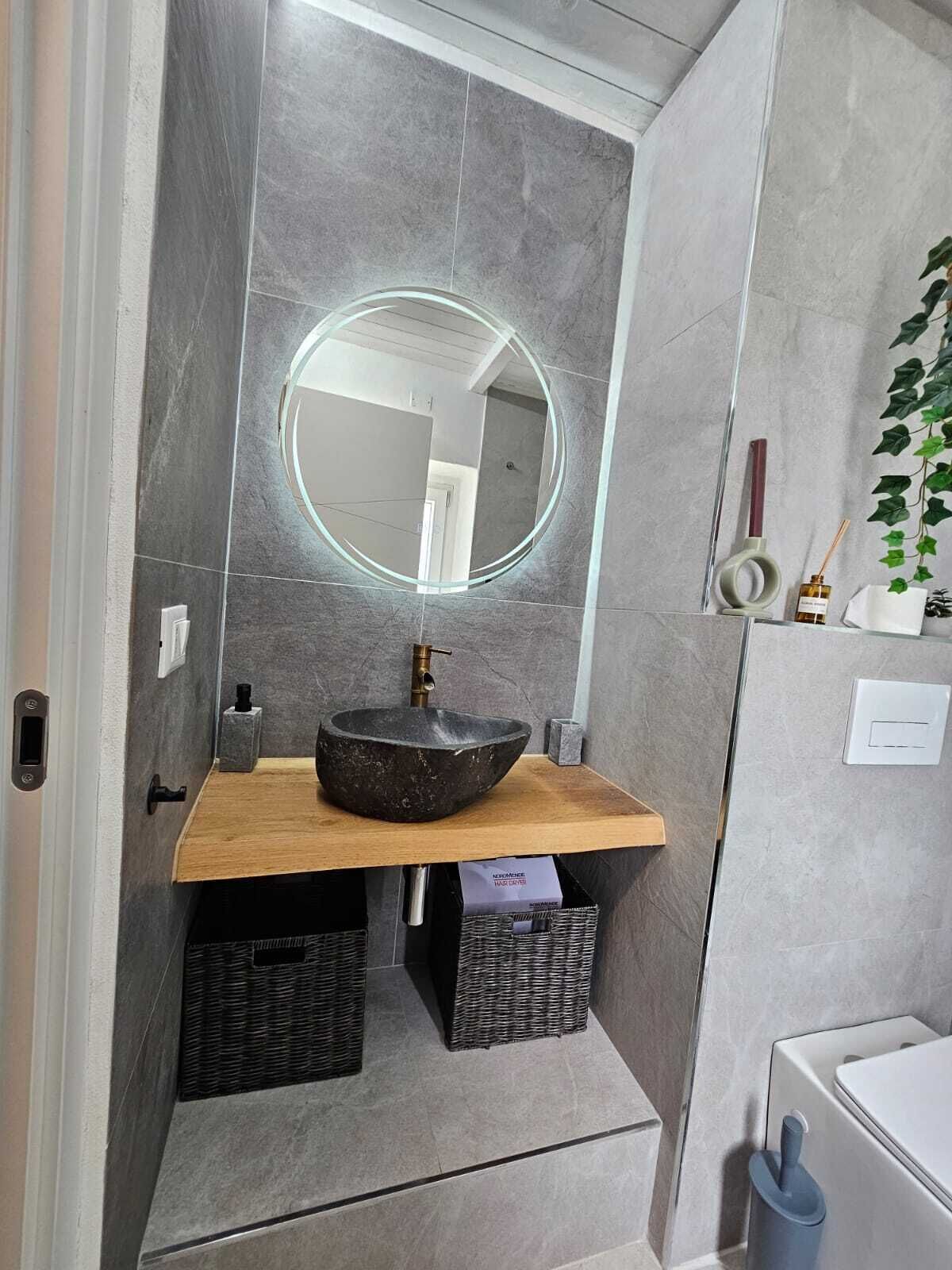 A modern bathroom with a round mirror, a stone sink, wicker baskets, and a toilet.