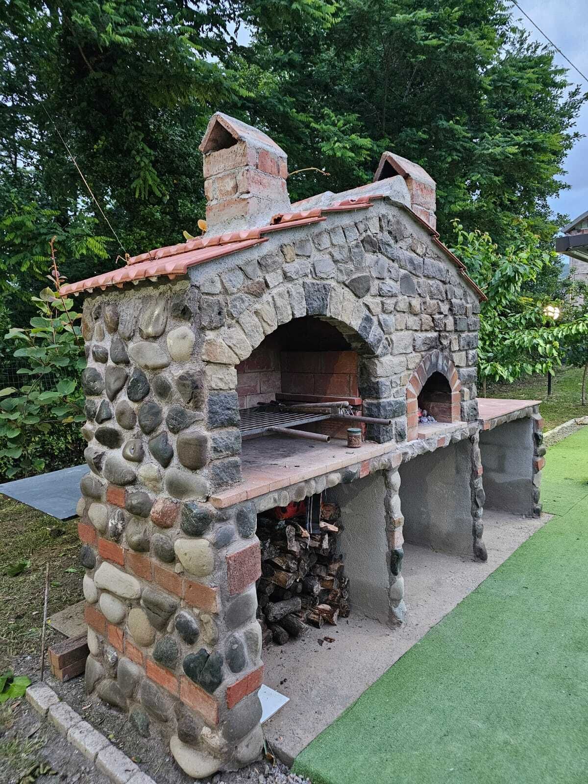 A stone-built outdoor oven with a brick chimney, surrounded by greenery.