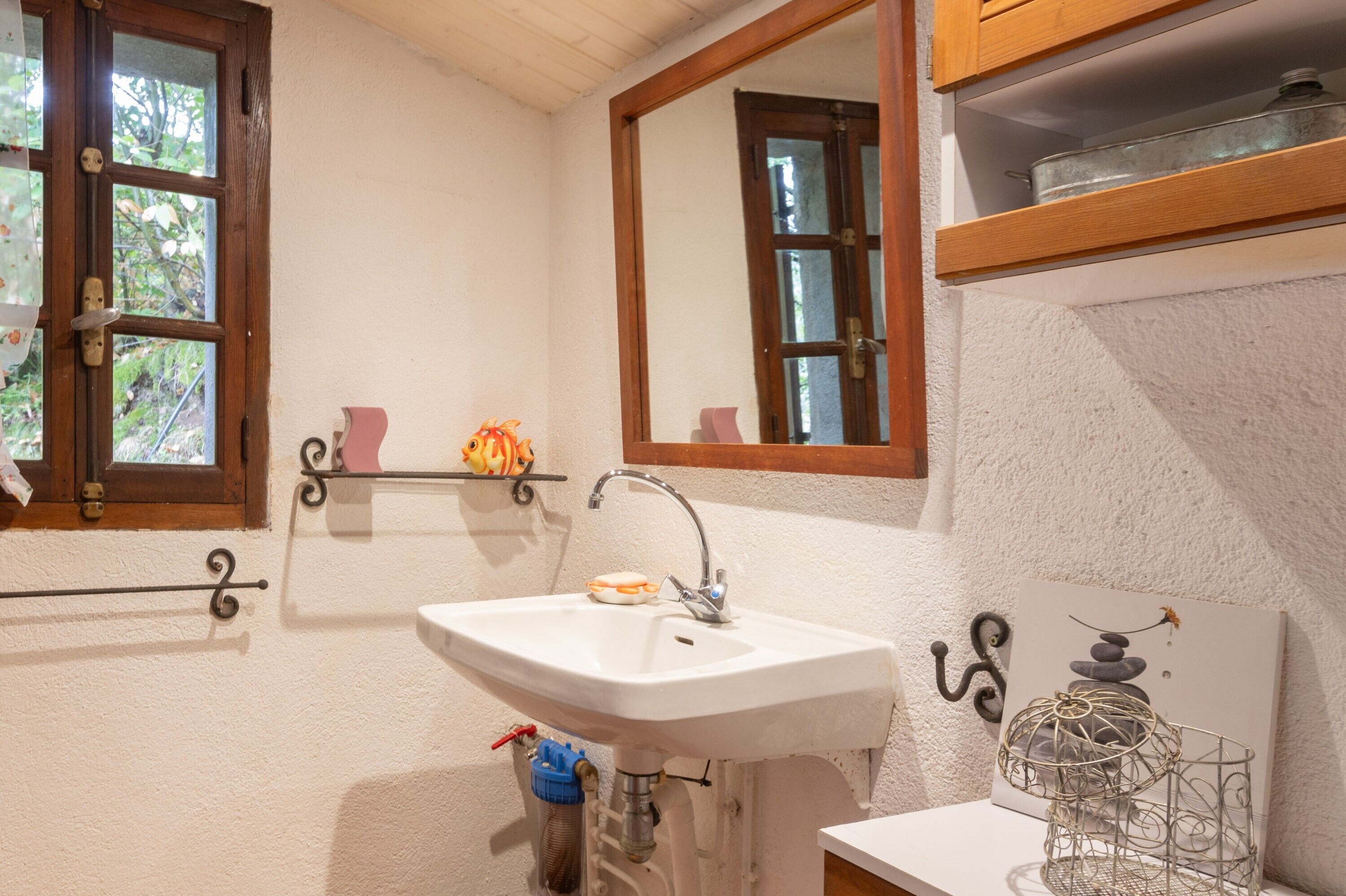 Un baño con lavamanos blanco, un espejo, una ventana y una repisa con objetos decorativos.
