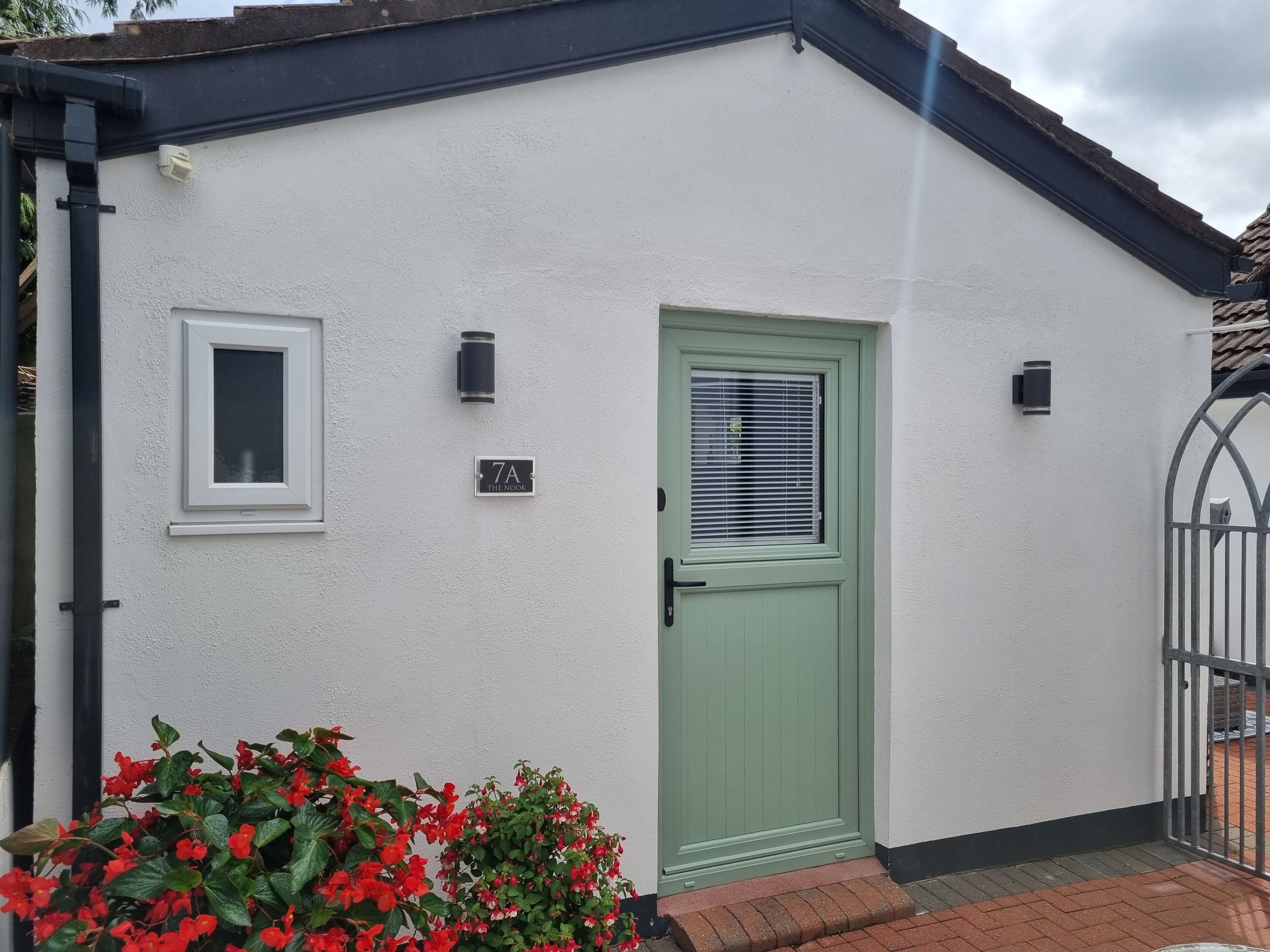 A white building with a green door, a small window, and a number plate reading "7A THE ROOR".